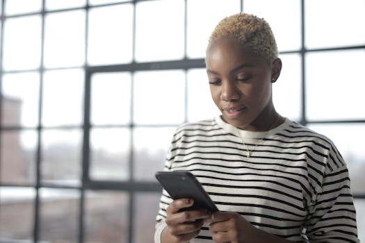 Young African American woman texting on smartphone by a window with natural light.